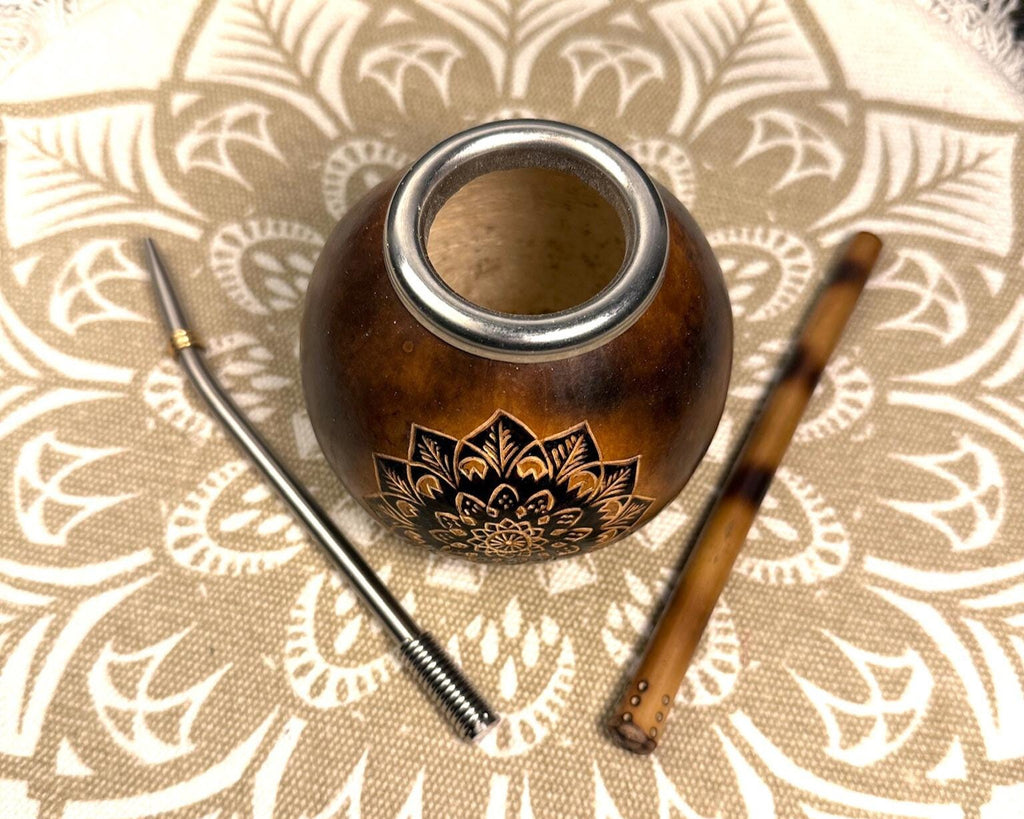 A small brown wooden mate pot with a silver rim and a black lotus engraving, shown with a metal bombilla straw and a brown wooden siphon on a beige patterned textile background.
