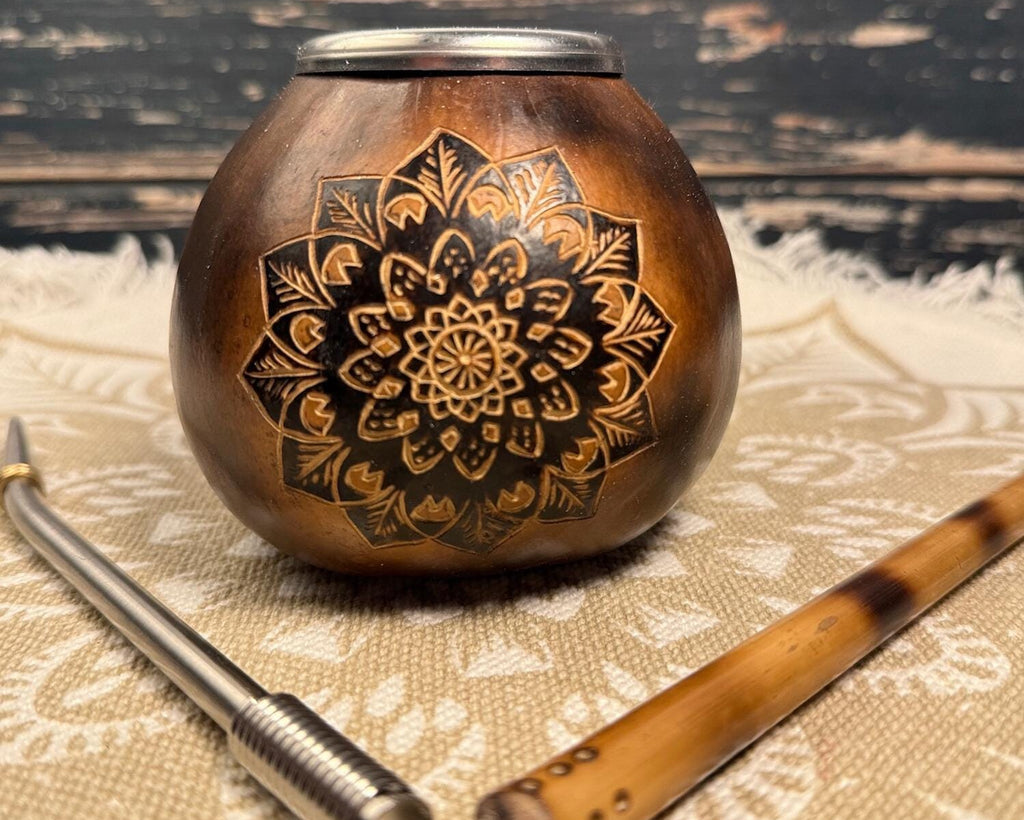 A round wooden mate pot with a carved black mandala design on the front, shown with a metal bombilla and a wooden chancla on a patterned beige cloth against a dark rustic wooden background.
