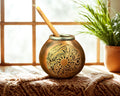 A round brass mate pot with intricate engraved patterns and a wooden spoon, set on a textured blanket in front of a window with a potted plant in the background.