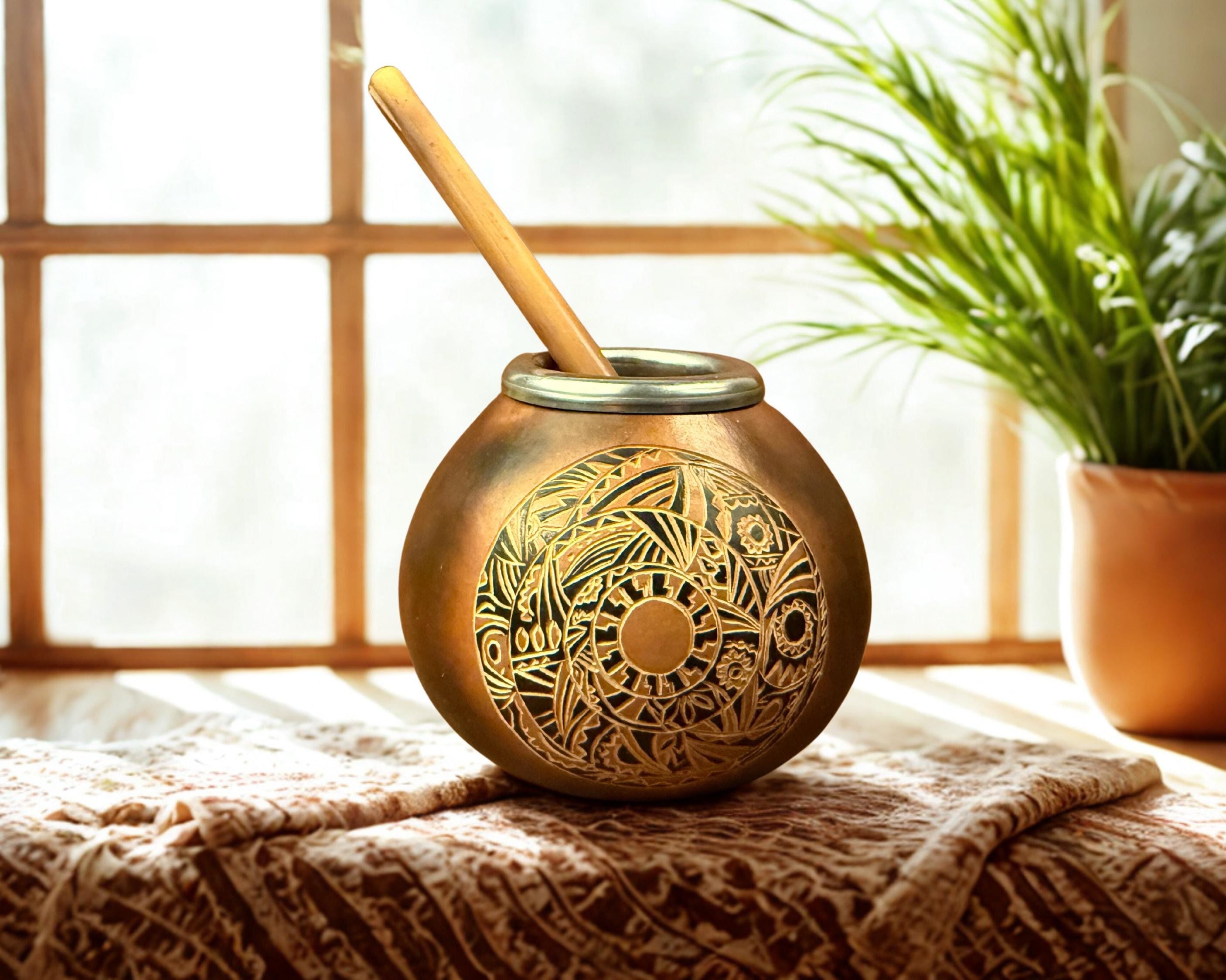 A round brass mate pot with intricate engraved patterns and a wooden spoon, set on a textured blanket in front of a window with a potted plant in the background.