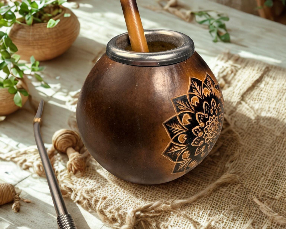 A traditional copper mate pot with a carved floral mandala design, filled with a wooden straw, set on a burlap cloth with a metalmate and plants in the background.