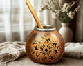 A copper mate cup with black floral engravings and a wooden spoon, styled on a table with a beige cloth and dried flowers in the background