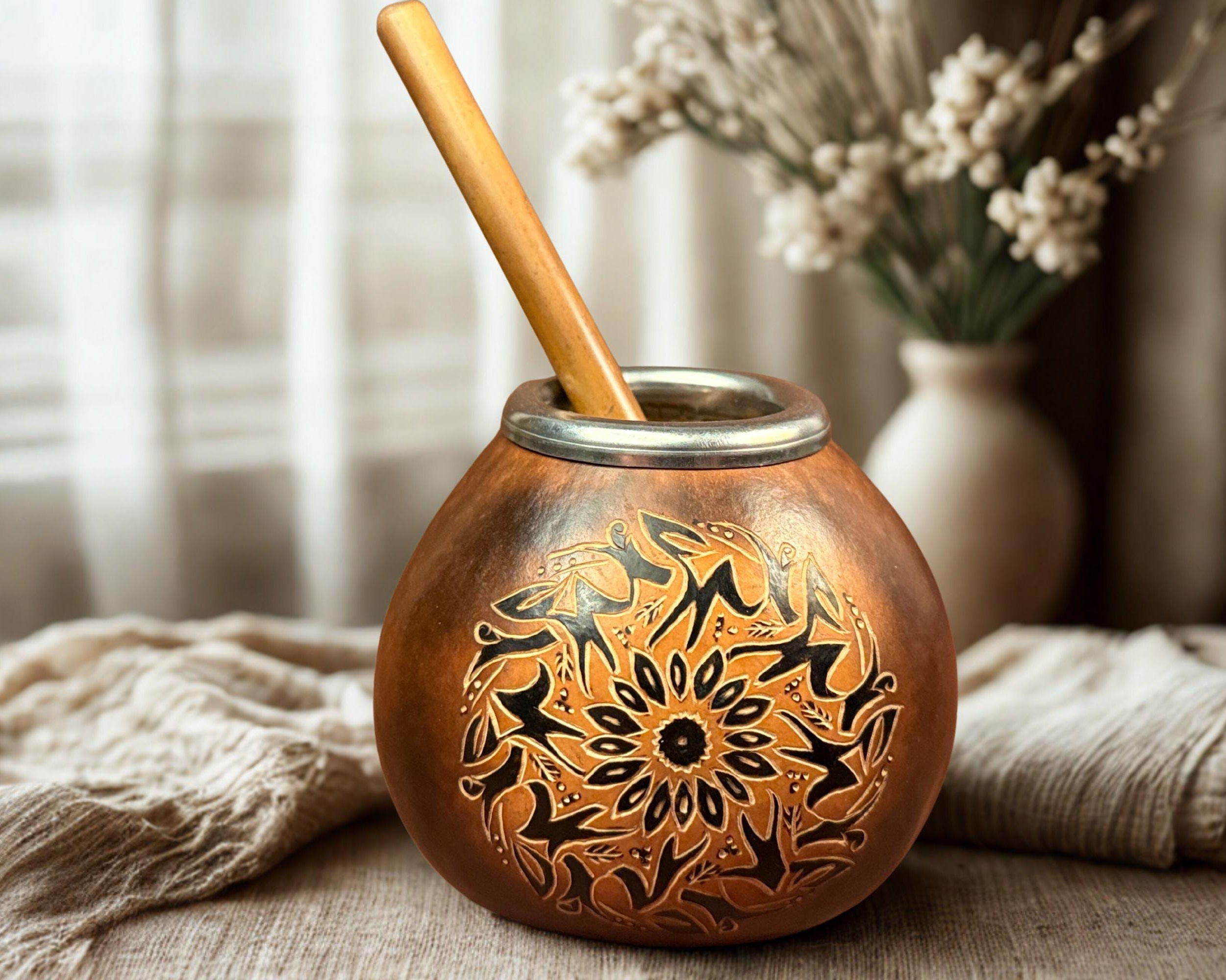 A copper mate cup with black floral engravings and a wooden spoon, styled on a table with a beige cloth and dried flowers in the background