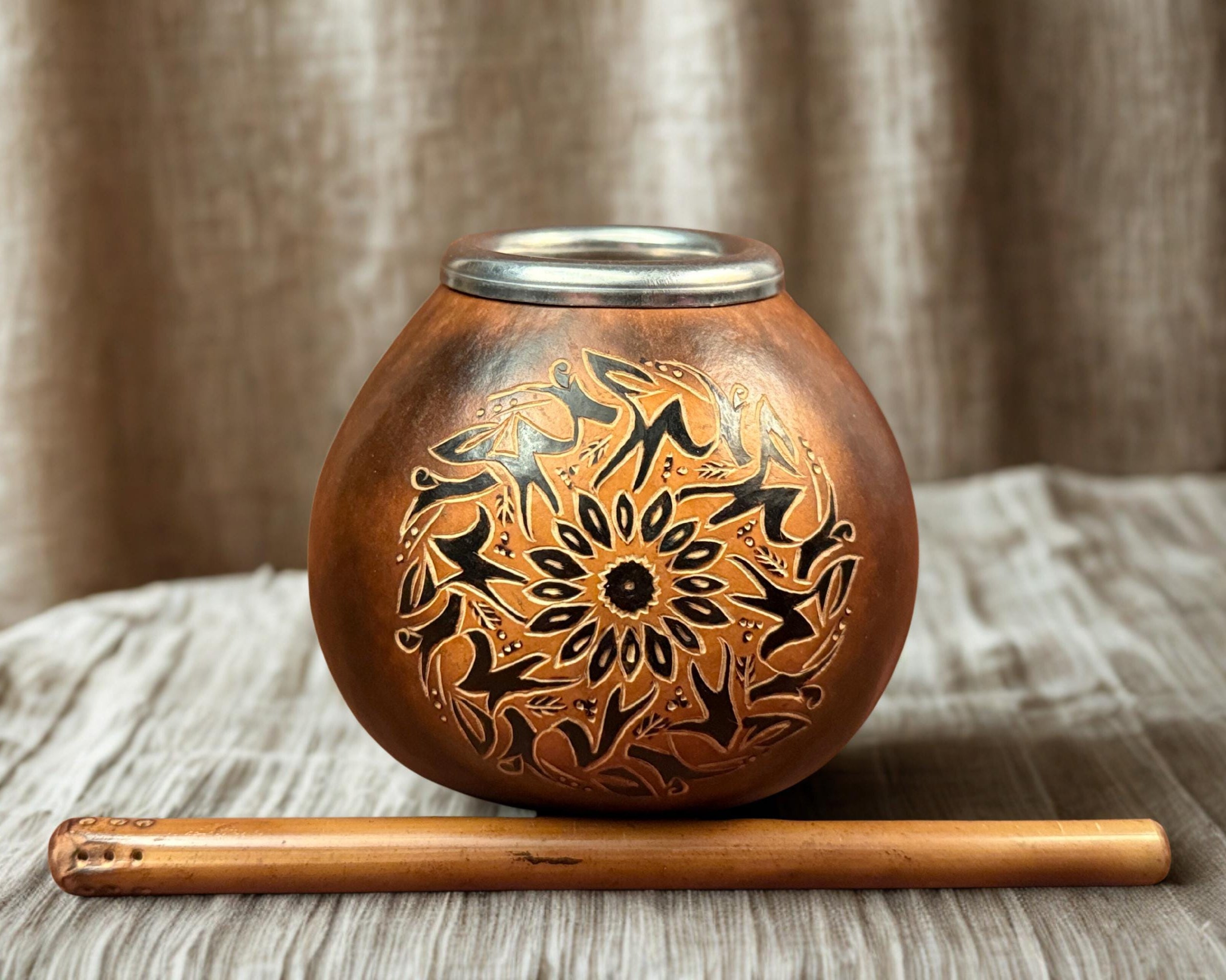 A round copper mate pot with an engraved black floral and geometric pattern, shown with a wooden flute in front of it on a textured beige fabric surface with a draped fabric background