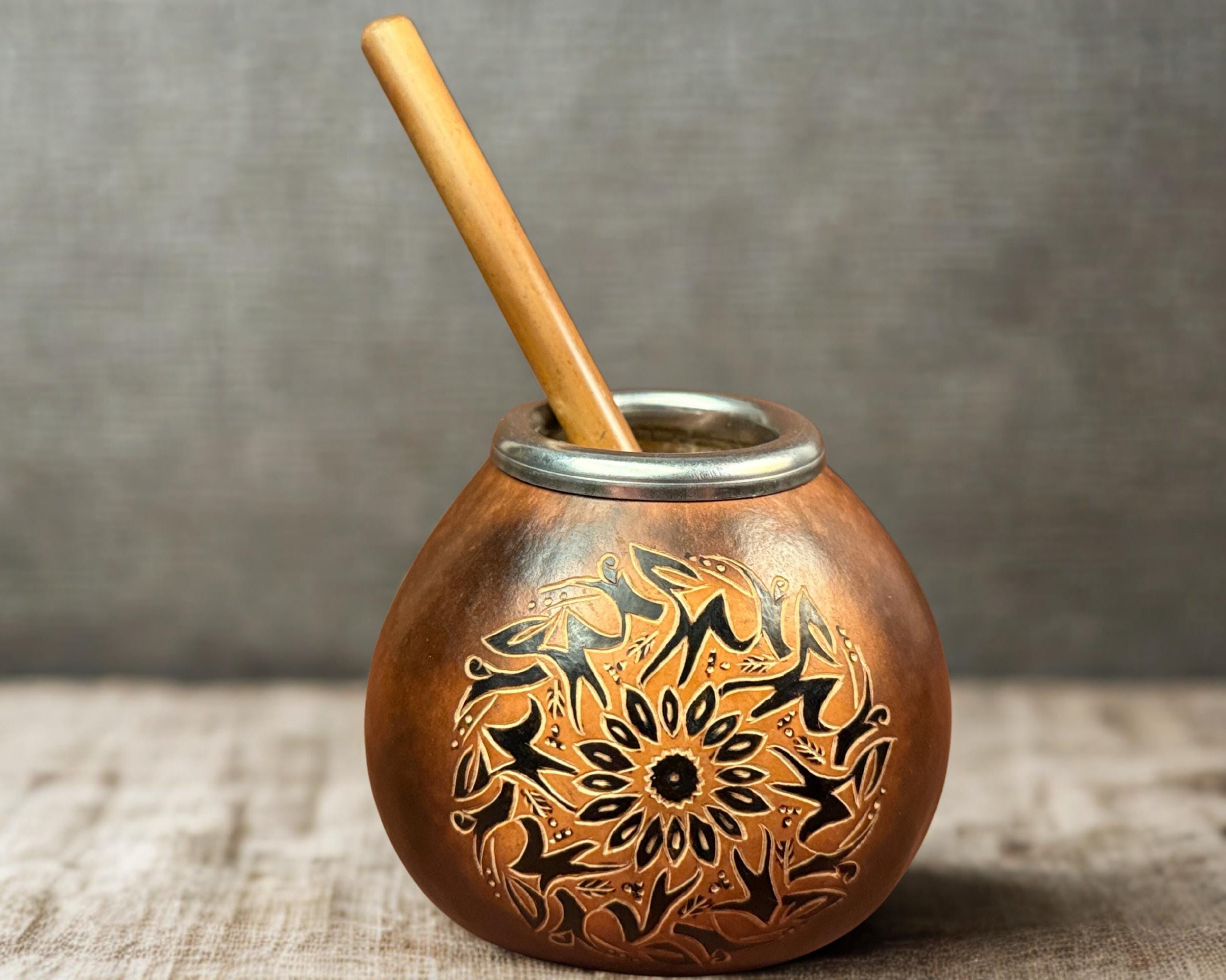 A traditional copper mate pot with an engraved floral mandala pattern, featuring a light brown wooden spoon resting inside, set on a rustic wooden surface against a textured gray background.