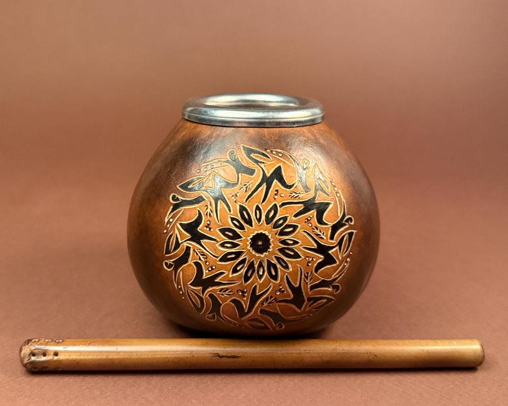 A small round copper-toned metal pot with a silver rim, decorated with black floral and geometric patterns, shown on a brown background with a wooden stick placed in front.