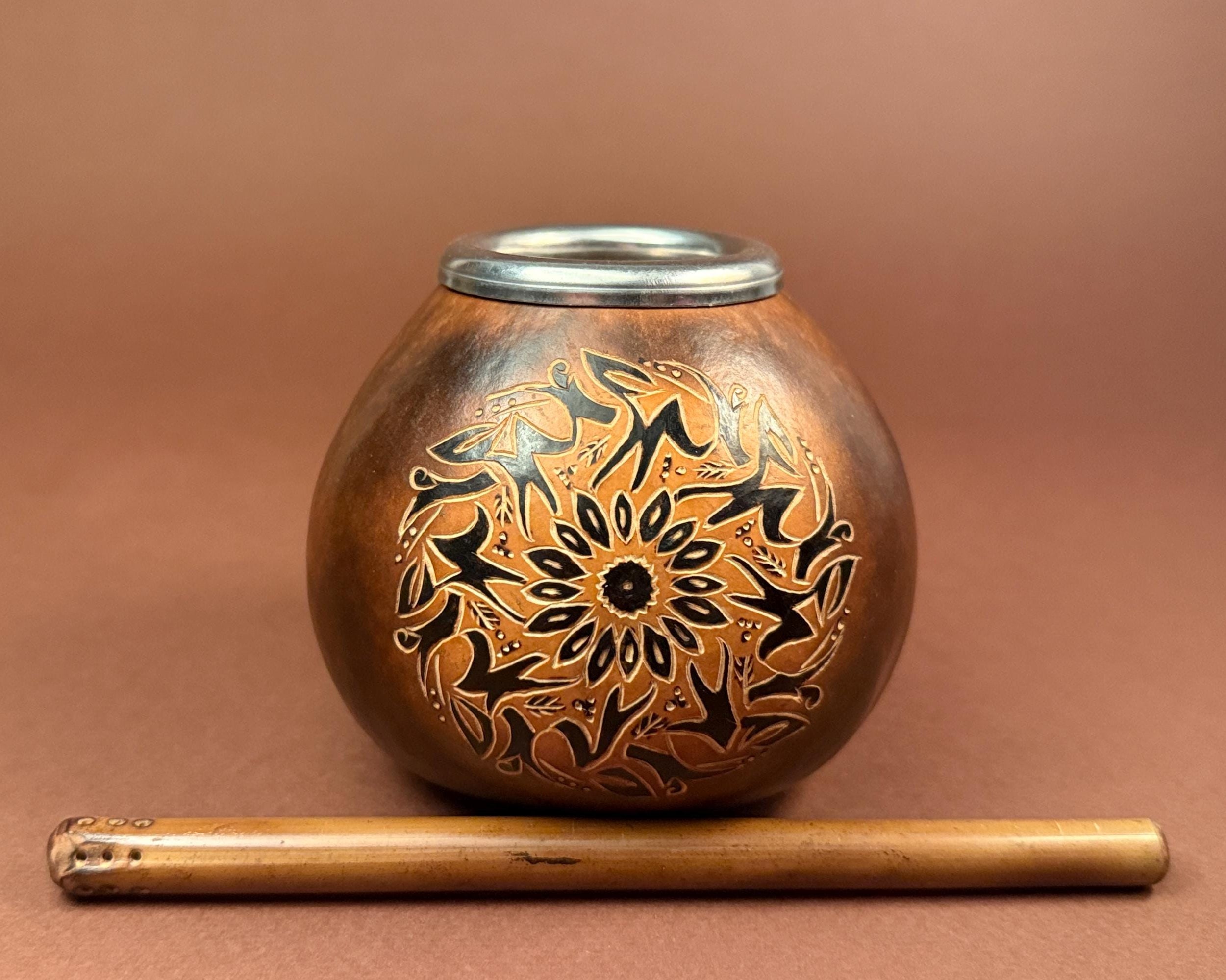 A small round copper-toned metal pot with a silver rim, decorated with black floral and geometric patterns, shown on a brown background with a wooden stick placed in front.
