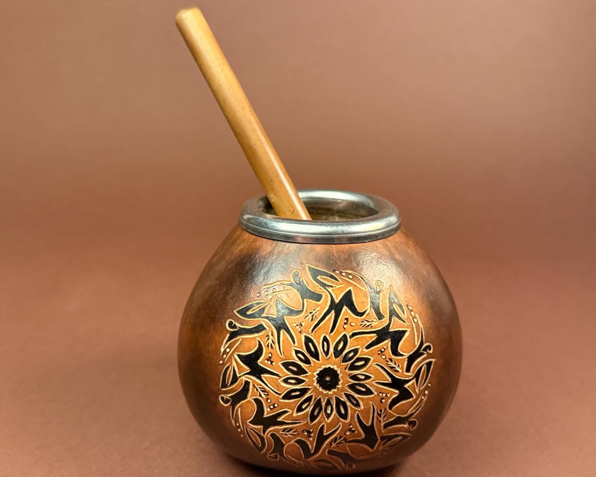 A copper mate pot with an engraved black floral mandala pattern and a light brown wooden straw, set against a solid brown background