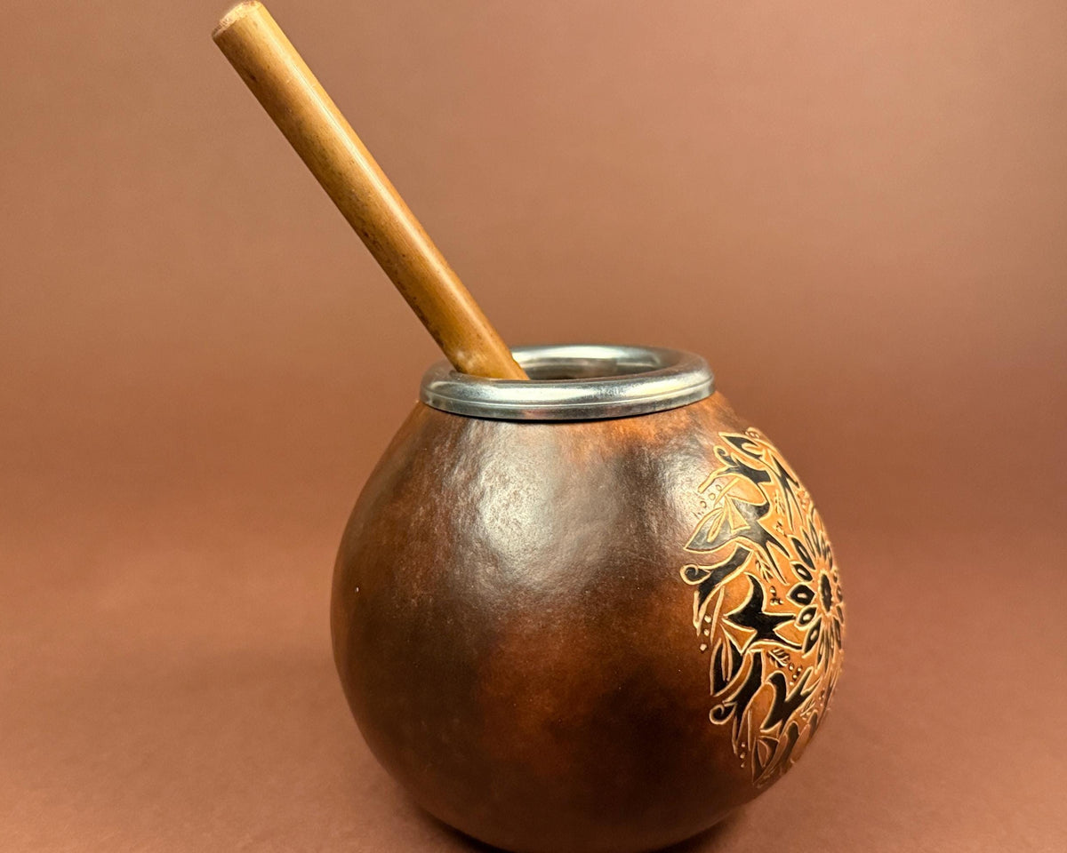 A brown handcrafted gourd with a wooden straw and silver rim, decorated with black and gold floral patterns, set against a solid brown background