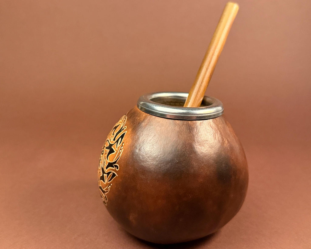 A brown leather mate bowl with a silver metal rim, a wooden straw, and a gold-colored floral engraving on the side, set against a solid brown background