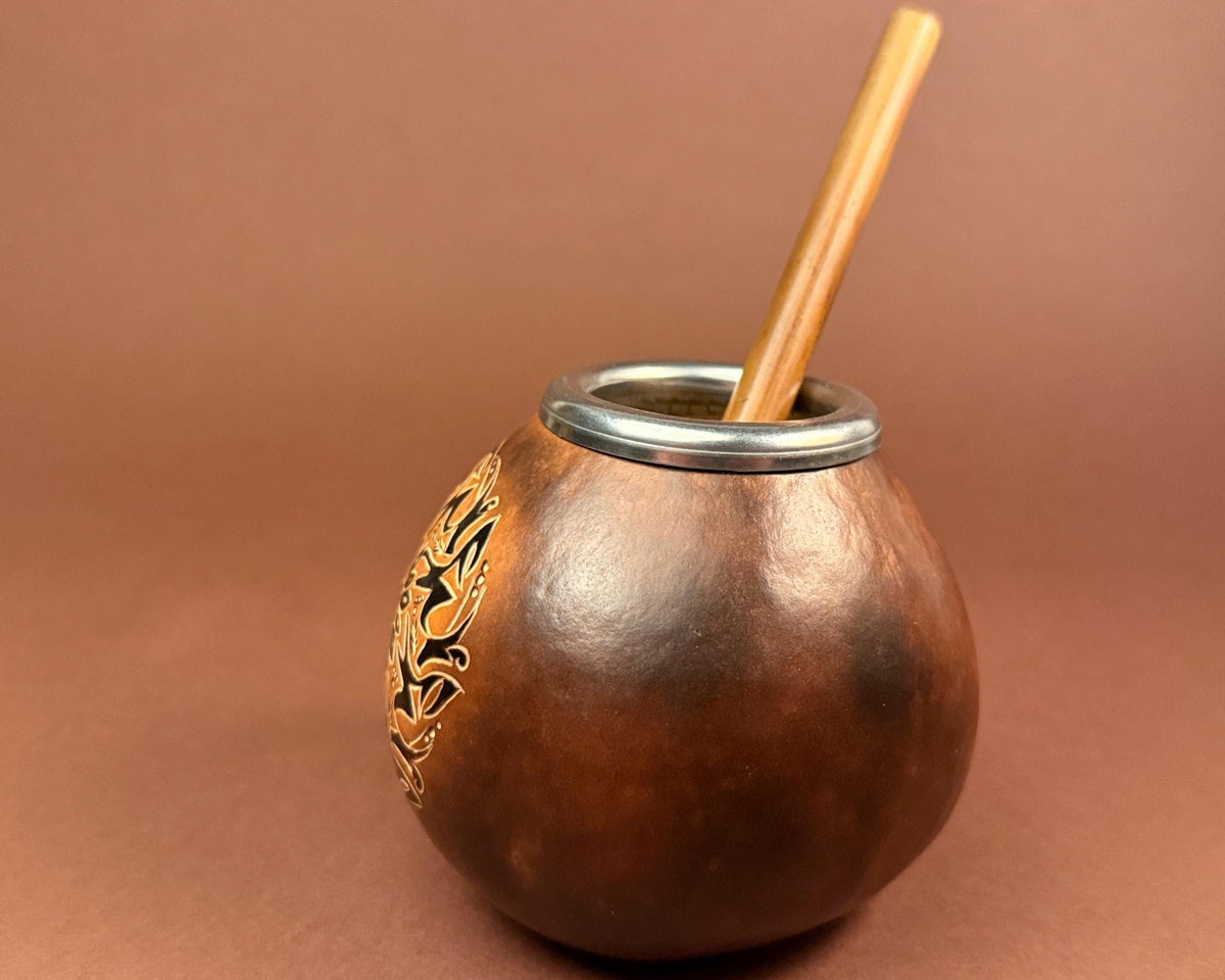 A brown leather mate bowl with a silver metal rim, a wooden straw, and a gold-colored floral engraving on the side, set against a solid brown background