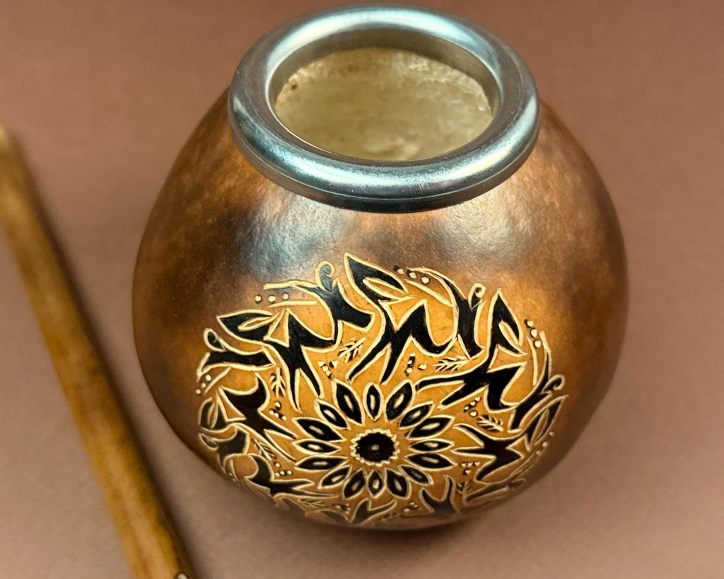 A handmade copper or brass mate pot with a silver rim and black hand-painted floral and geometric patterns, shown next to a wooden spoon on a brown background