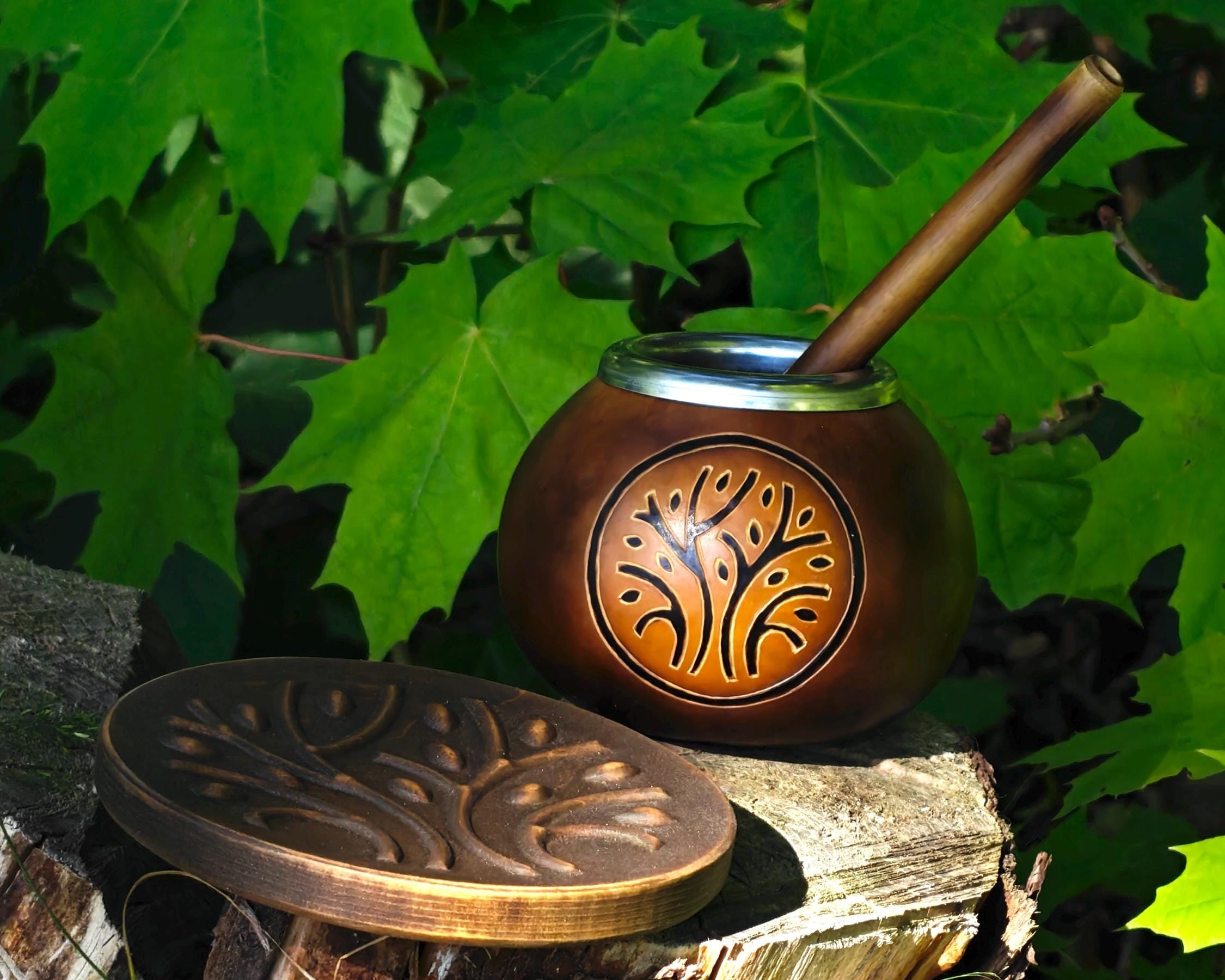 A brown wooden mate bomb with a carved tree design on the front, shown with its matching lid resting beside it on a tree stump outdoors, surrounded by green leaves