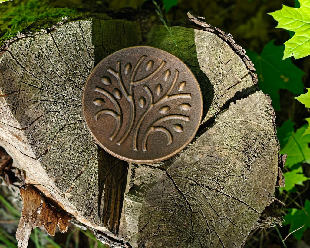 A round bronze-toned metal plaque with an embossed tree design, displayed on a cut tree stump surrounded by green leaves outdoors