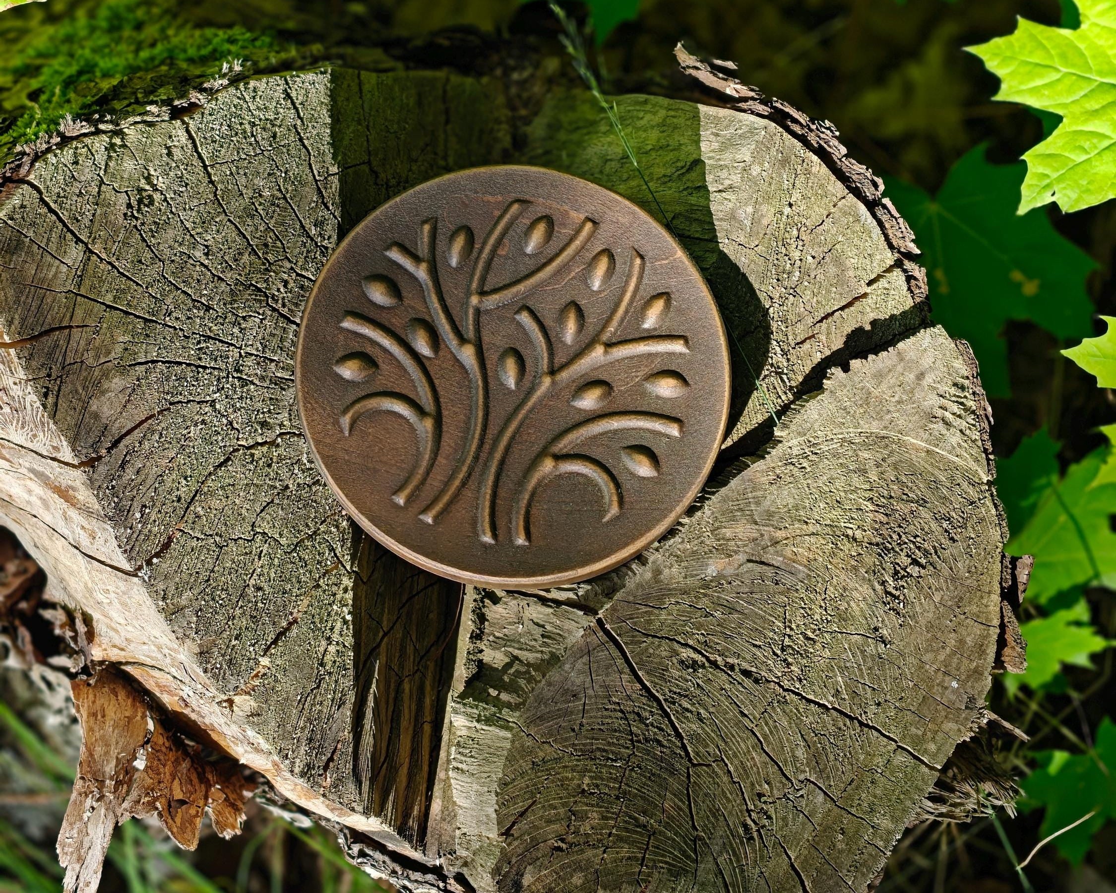 A round bronze-toned metal plaque with an embossed tree design, displayed on a cut tree stump surrounded by green leaves outdoors