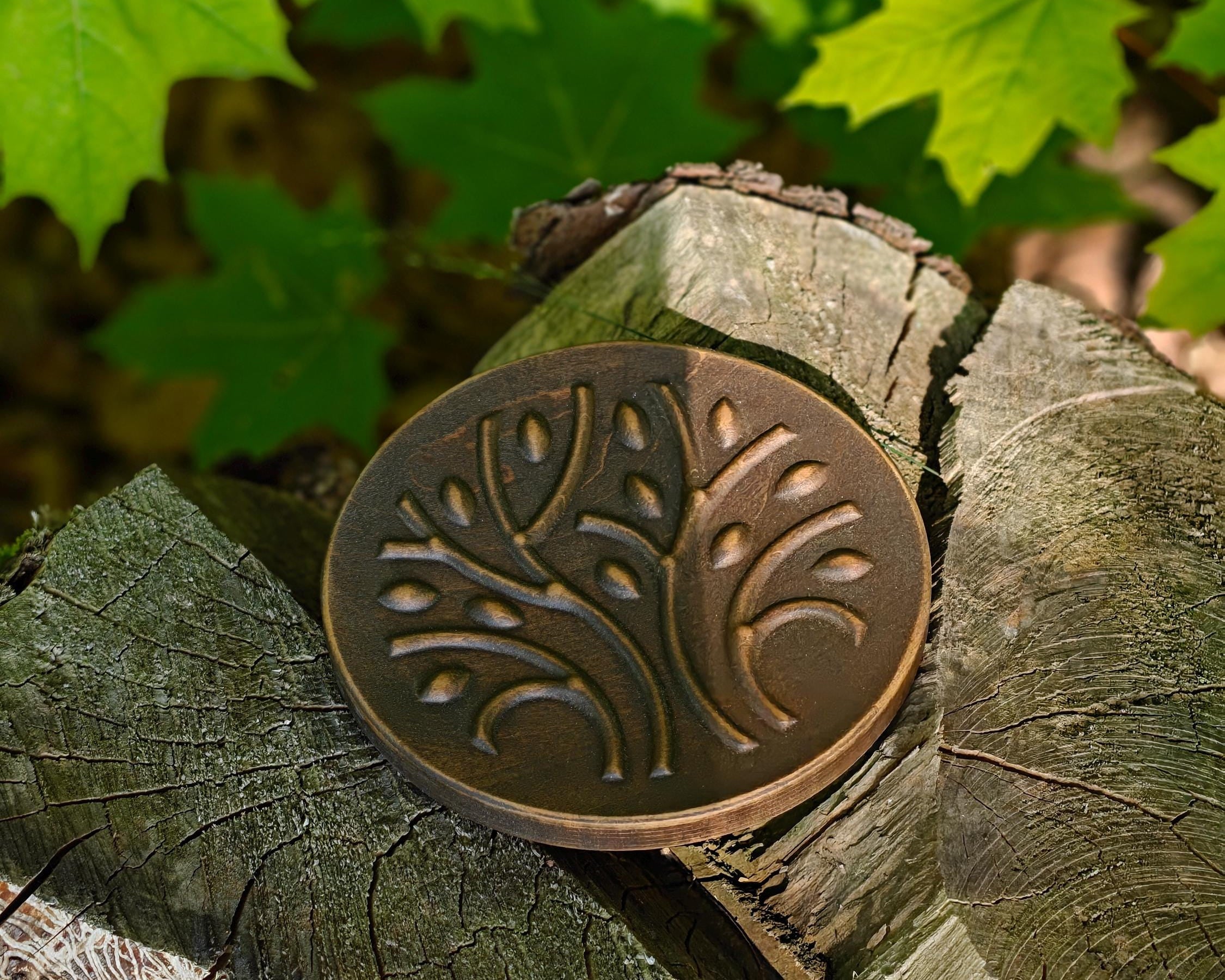 A round bronze metal coin or medallion with an embossed tree design, resting on a cut tree stump with green maple leaves in the background