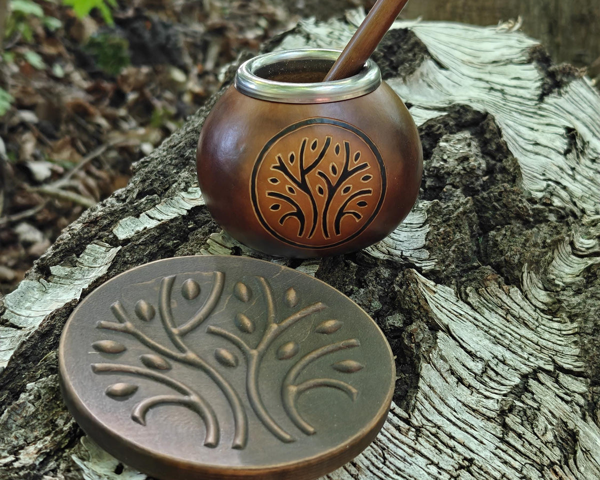 A copper mate pot with a wooden straw sits on a tree stump outdoors, with its matching round lid placed in front. The copper has an engraved tree design, and the background shows natural forest ground and wood