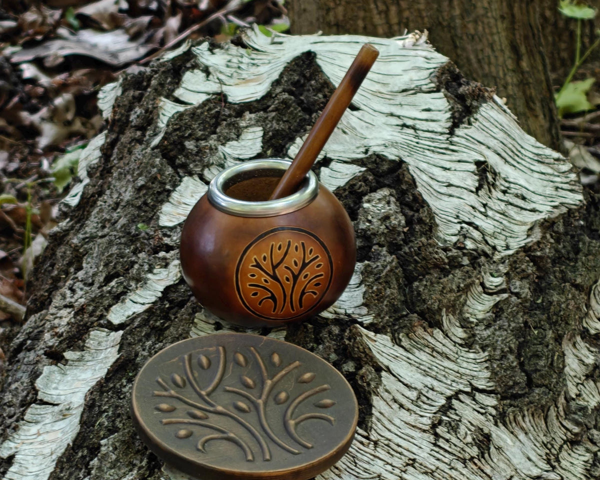 A small copper-toned mate pot with a wooden spoon and a matching round lid, both engraved with a tree motif, displayed on a cut tree stump outdoors.