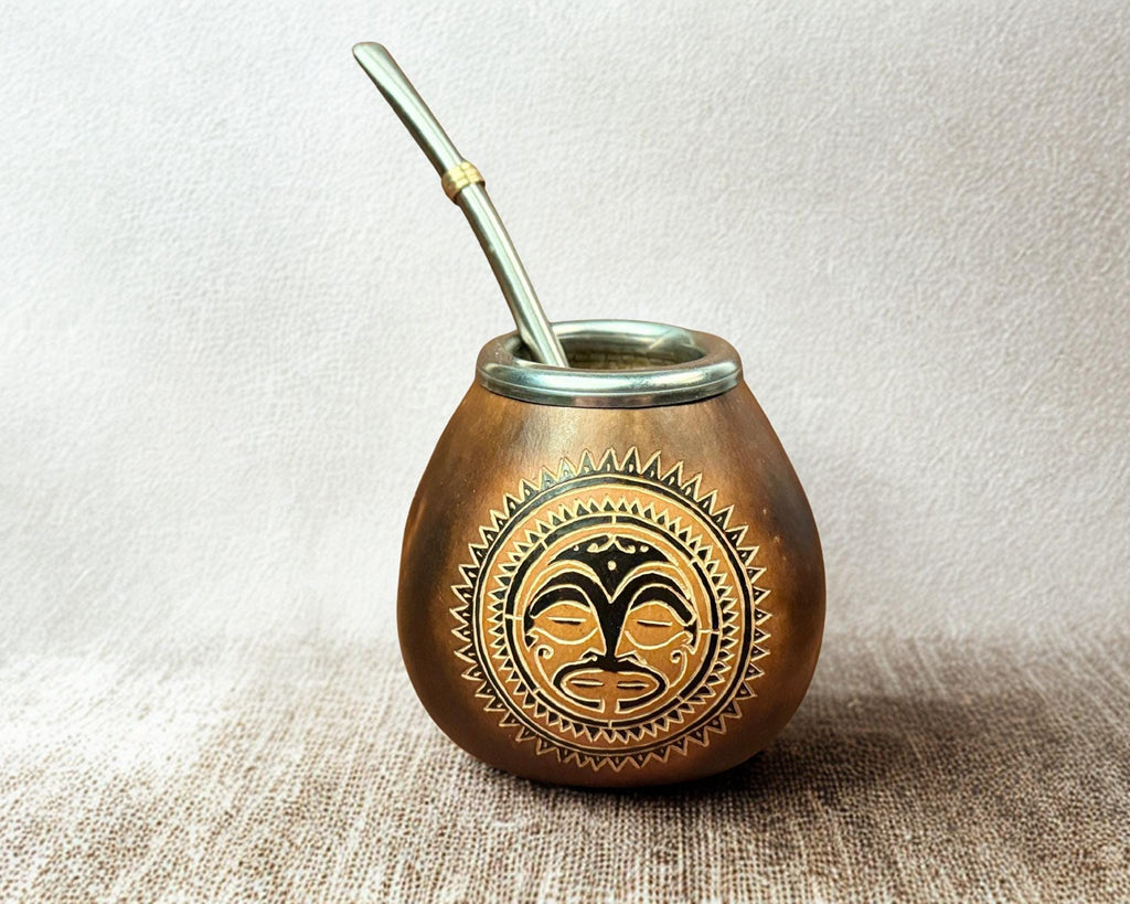 A small copper-colored metal yerba mate cup with a matching metal straw, featuring a black and gold tribal face design on the front, set against a neutral textured background