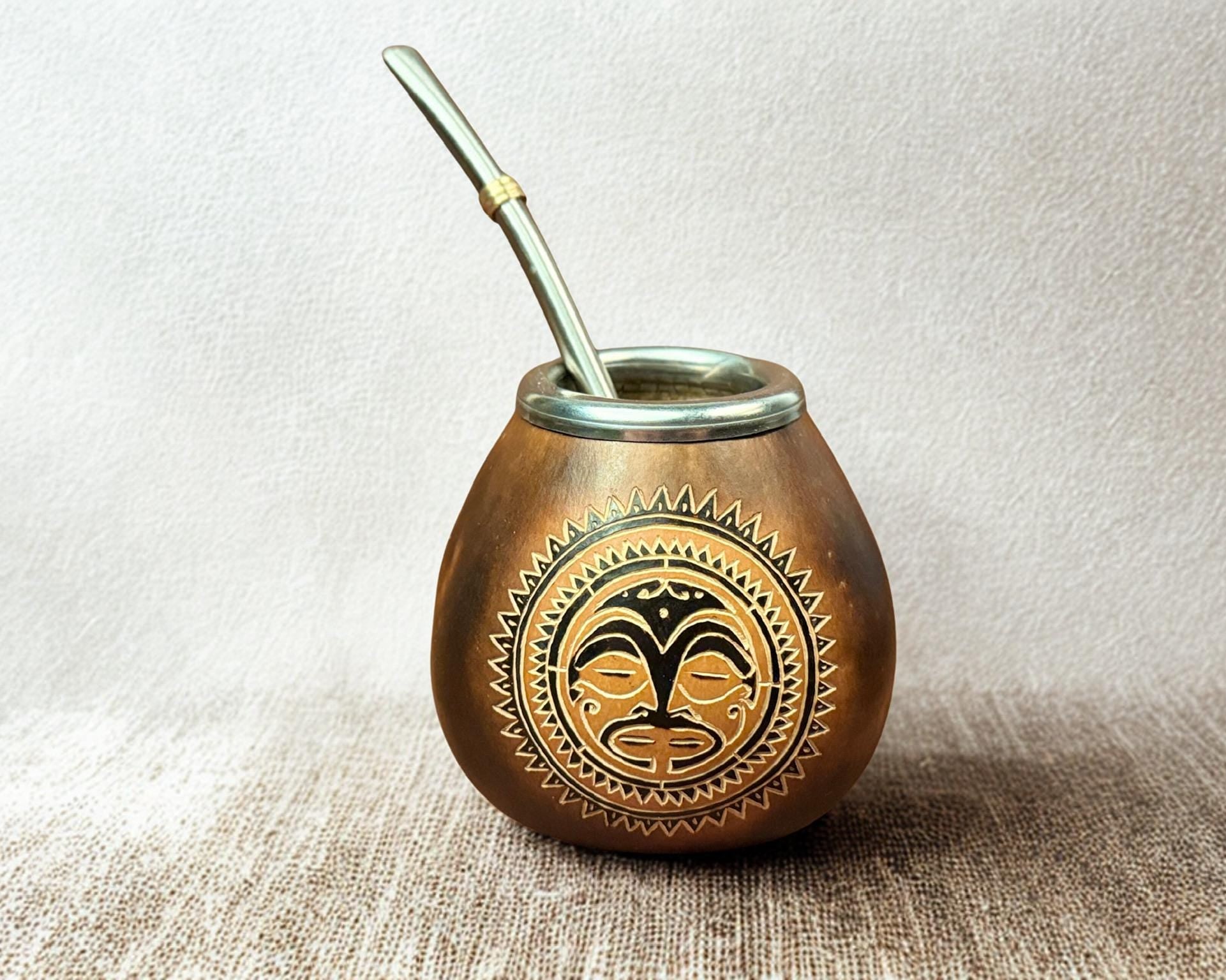 A small copper-colored metal yerba mate cup with a matching metal straw, featuring a black and gold tribal face design on the front, set against a neutral textured background