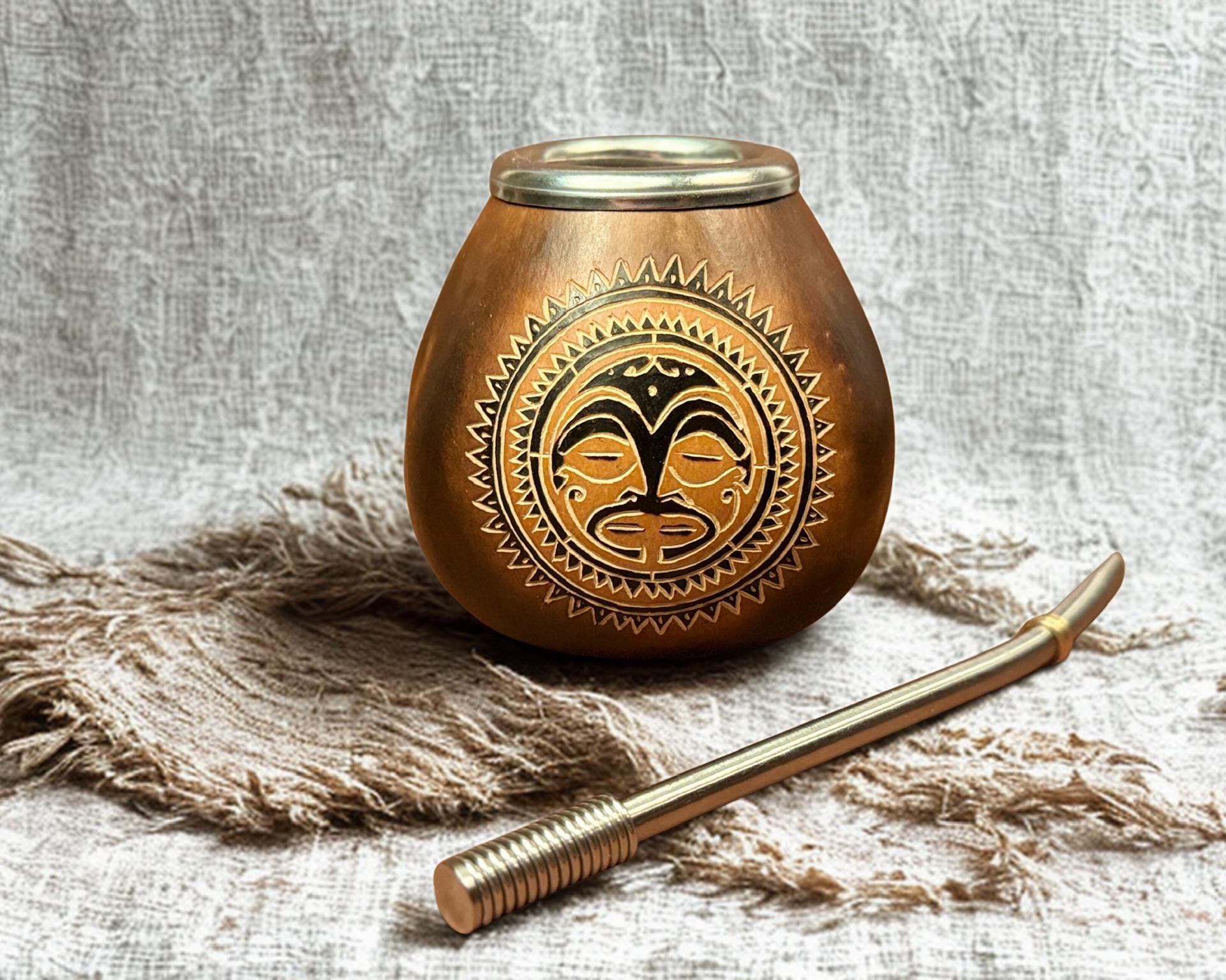 A brass or bronze ceremonial cup with a carved face motif on the front, shown with a matching metal scoop on a textured beige fabric background