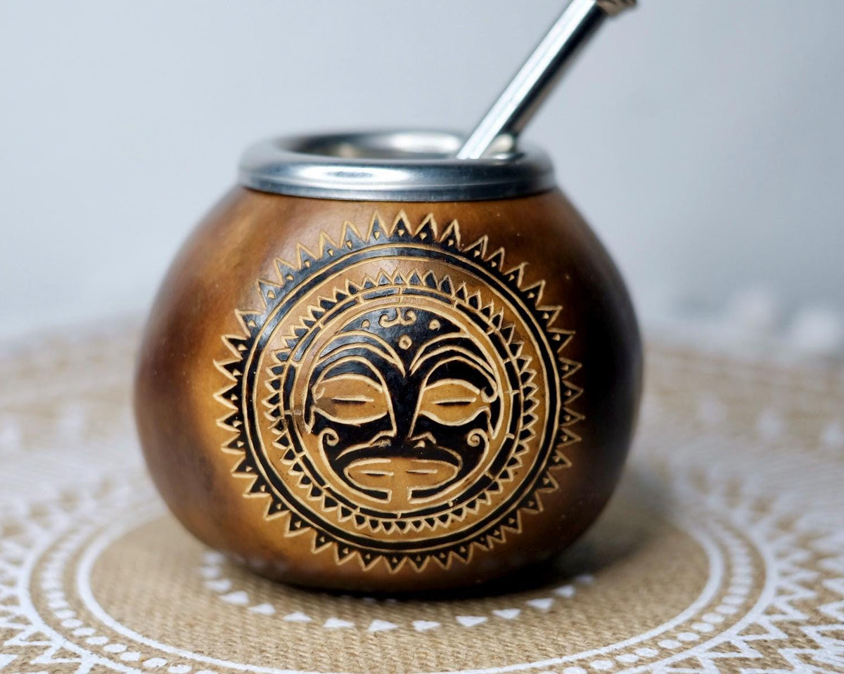 A brown wooden mate cup with a silver metal rim and straw, decorated with a black and gold tribal mask design on the front, resting on a lace doily