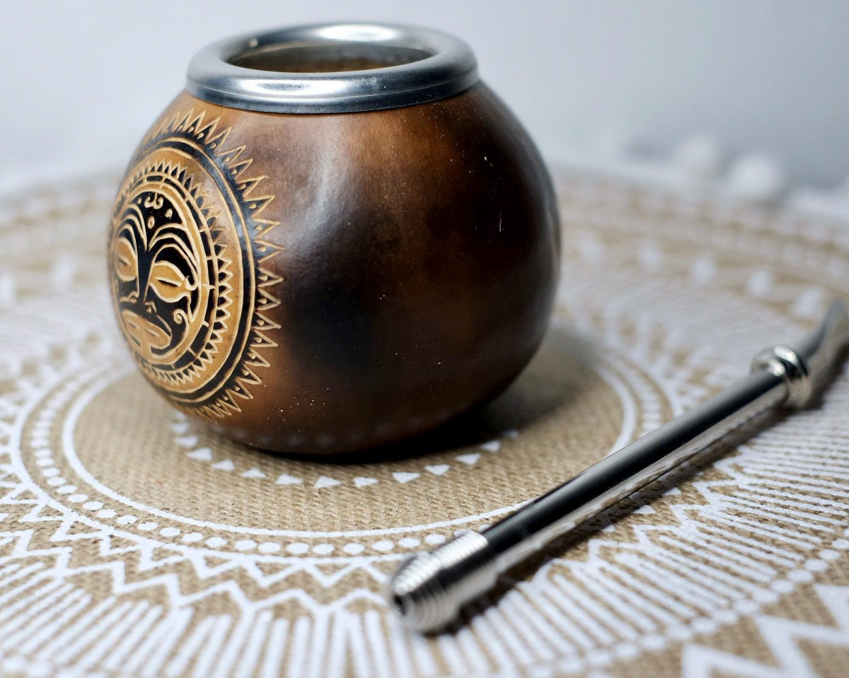 A traditional brown wooden mate pot with gold and black decorative engravings, shown next to a metal bombilla on a patterned beige placemat