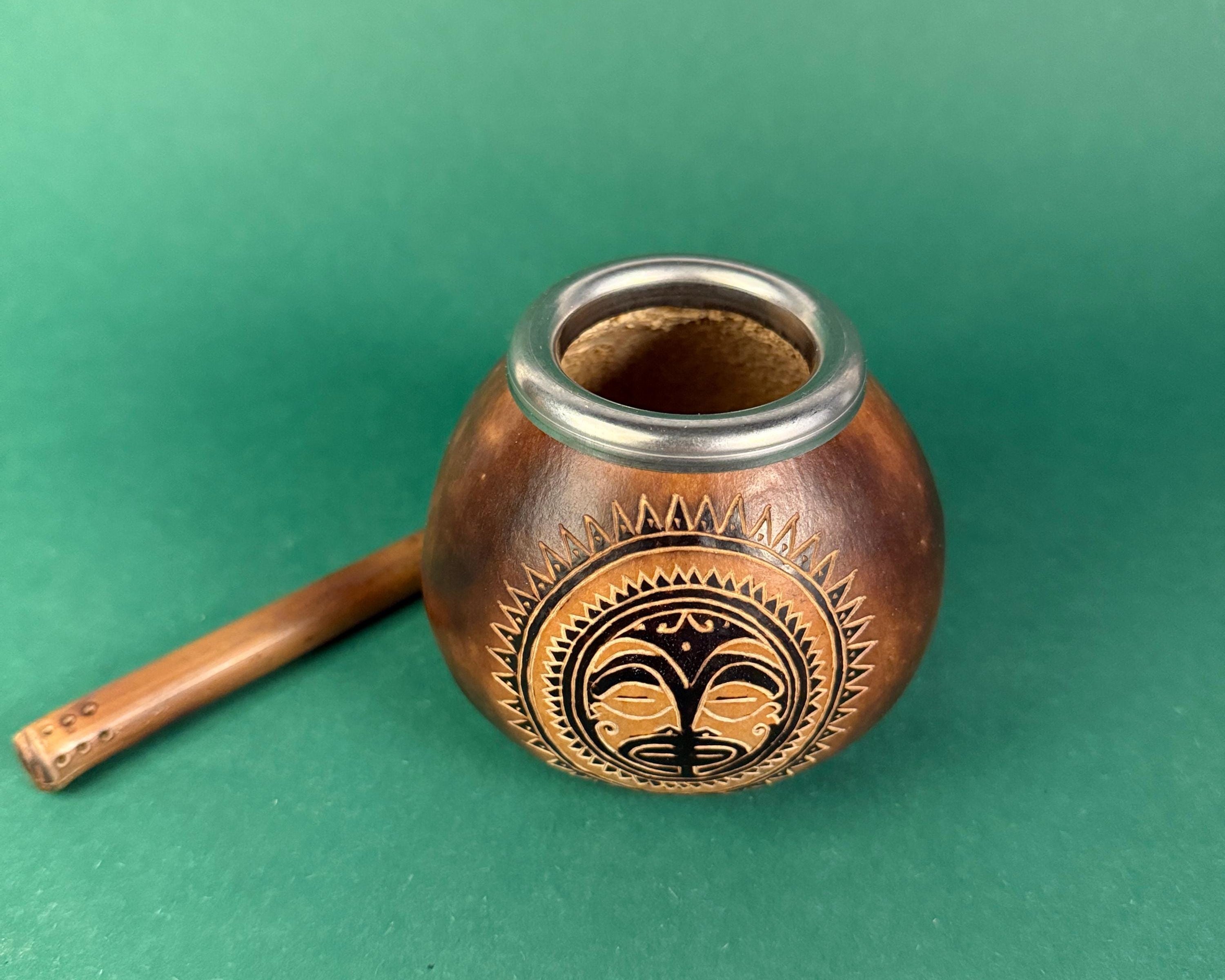 A small handcrafted wooden smoking bowl with a matching wooden pipe, featuring a carved black sun-and-moon style emblem on the front, set against a solid green background.