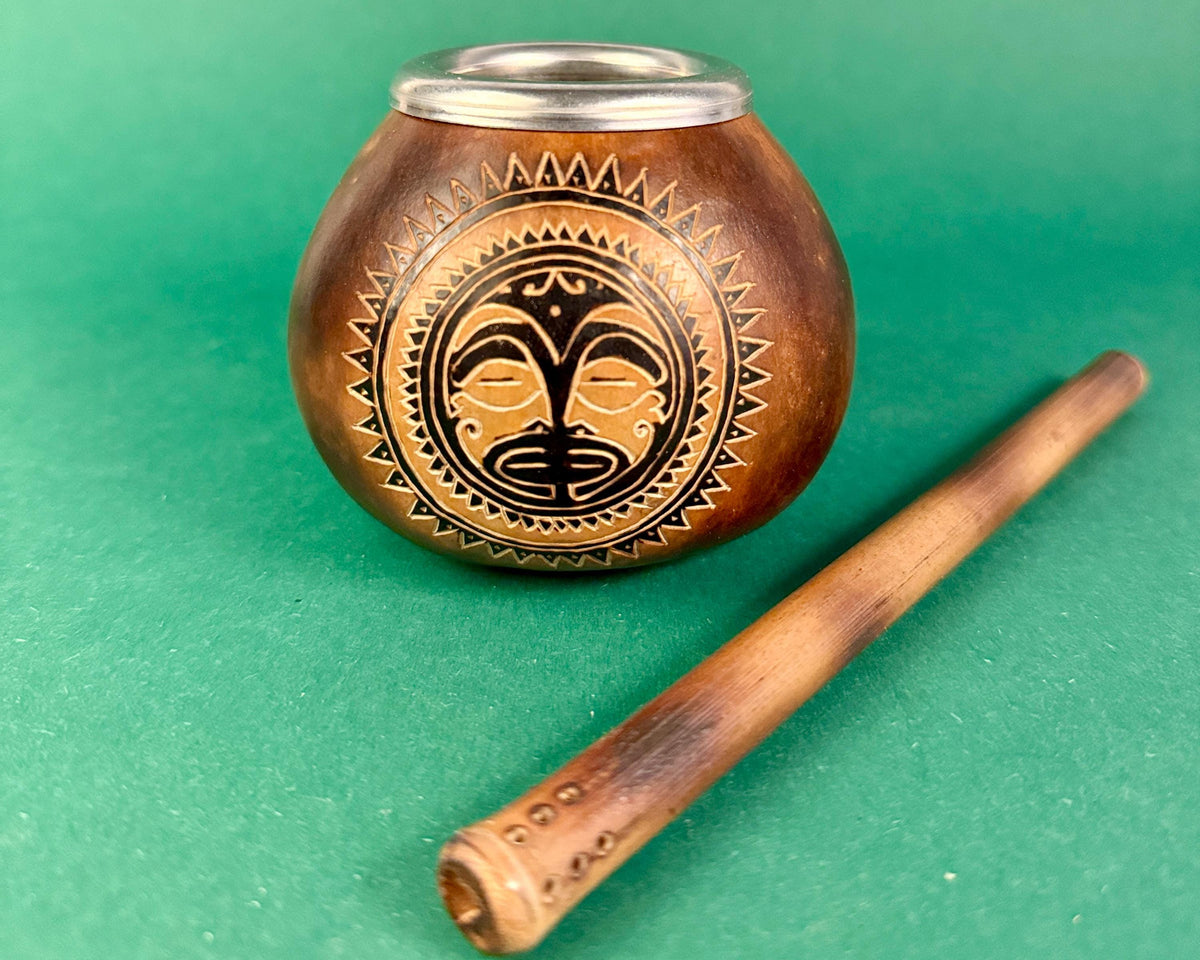 A small carved wooden smoking vessel with a silver metal rim and a black-and-brown tribal face motif on the front, shown with a wooden pipe on a green background