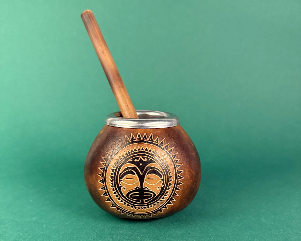 A small handcrafted wooden mate pot with a wooden spoon, featuring an engraved African-style face design on the front, set against a solid green background