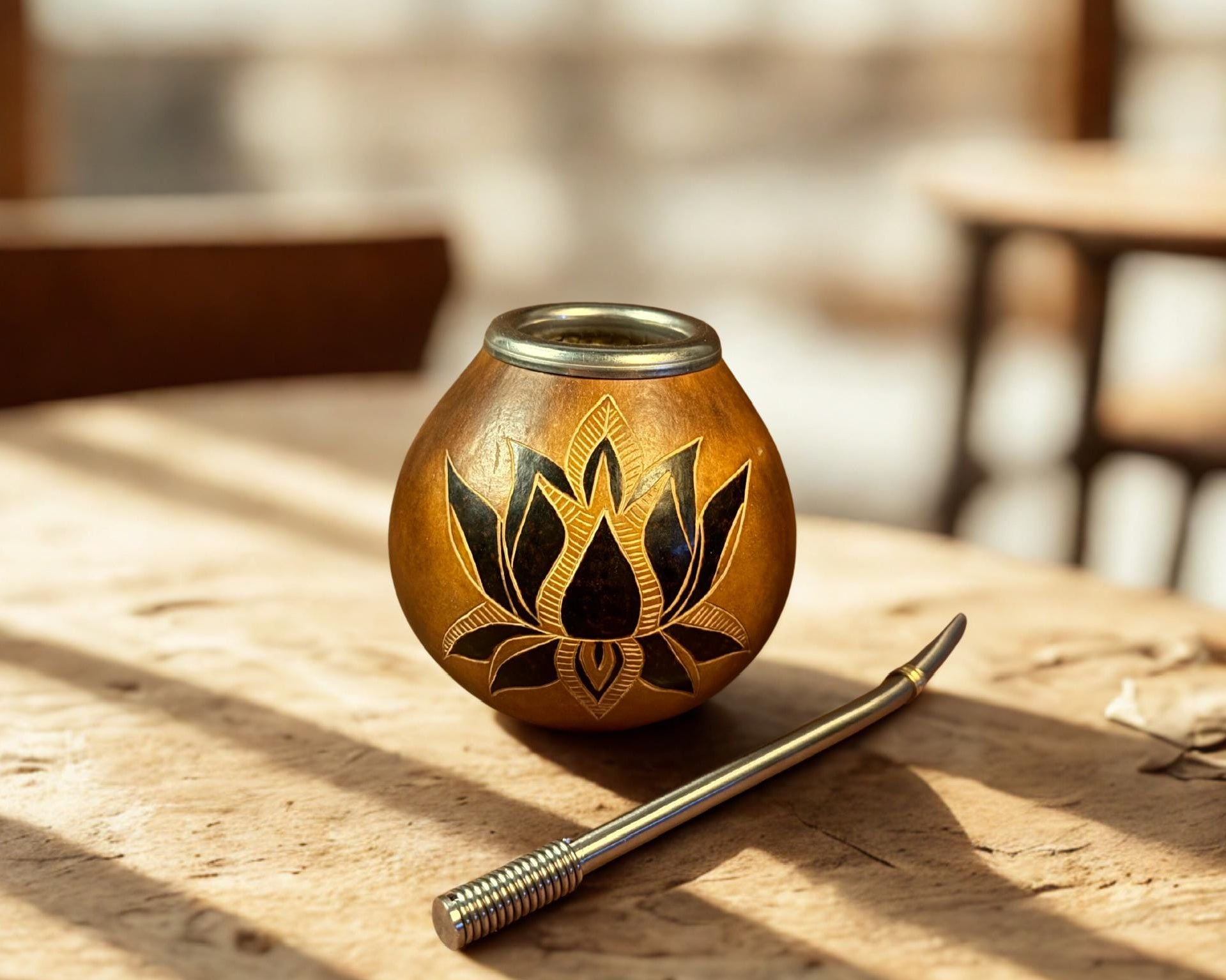 cup with a black lotus floral design, placed on a rustic wooden table next to a metal straw in a warm, sunlit setting