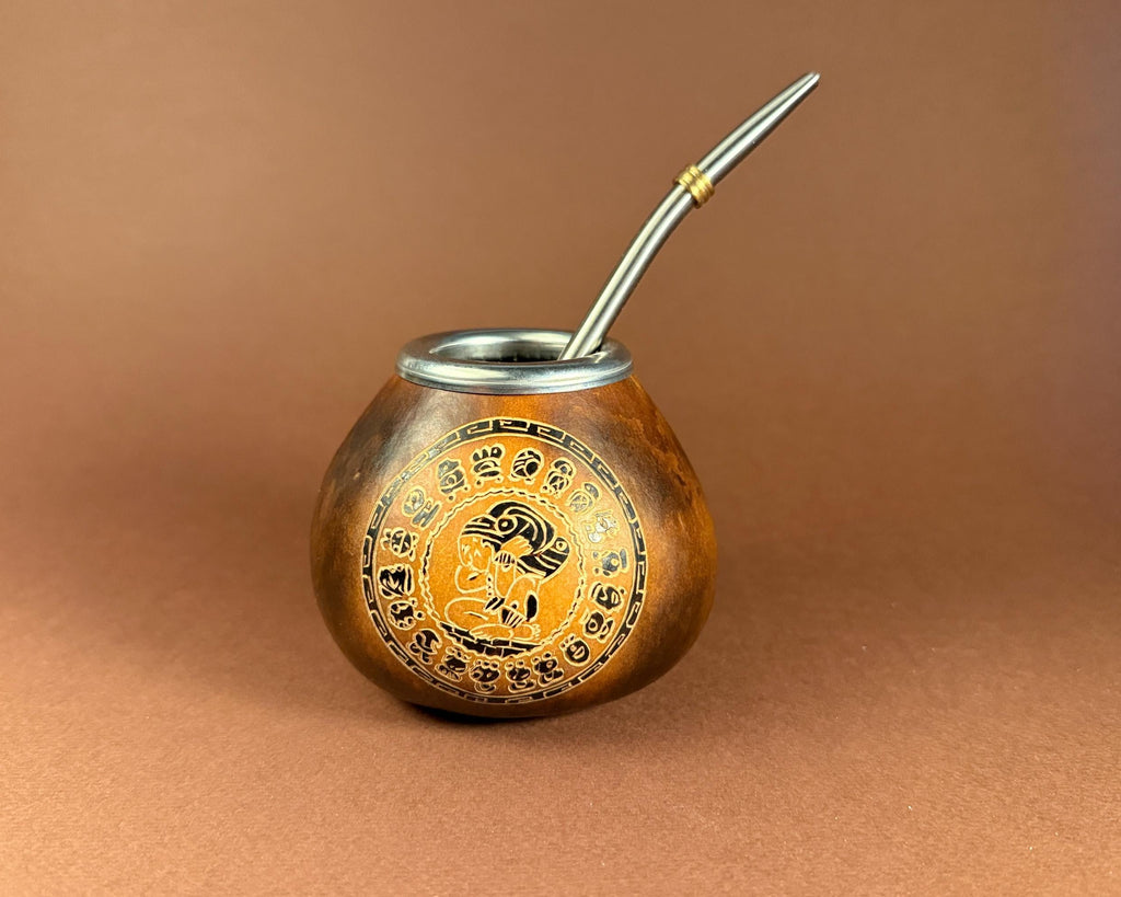 A traditional copper mate pot with a silver metal bombilla, featuring a circular gold and black decorative emblem on the front, set against a solid brown background.