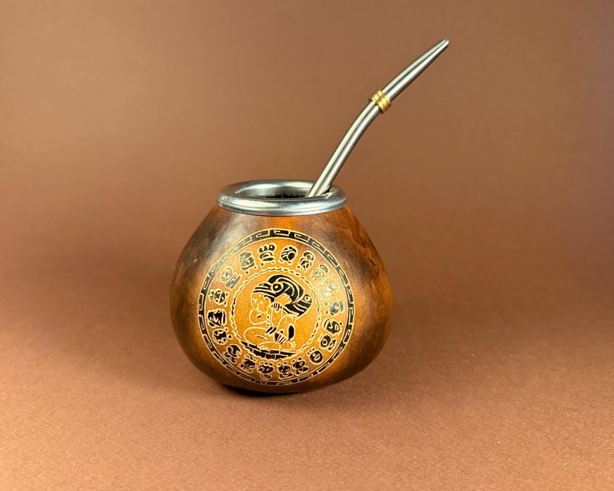 A traditional copper mate pot with a silver metal bombilla, featuring a circular gold and black decorative emblem on the front, set against a solid brown background.