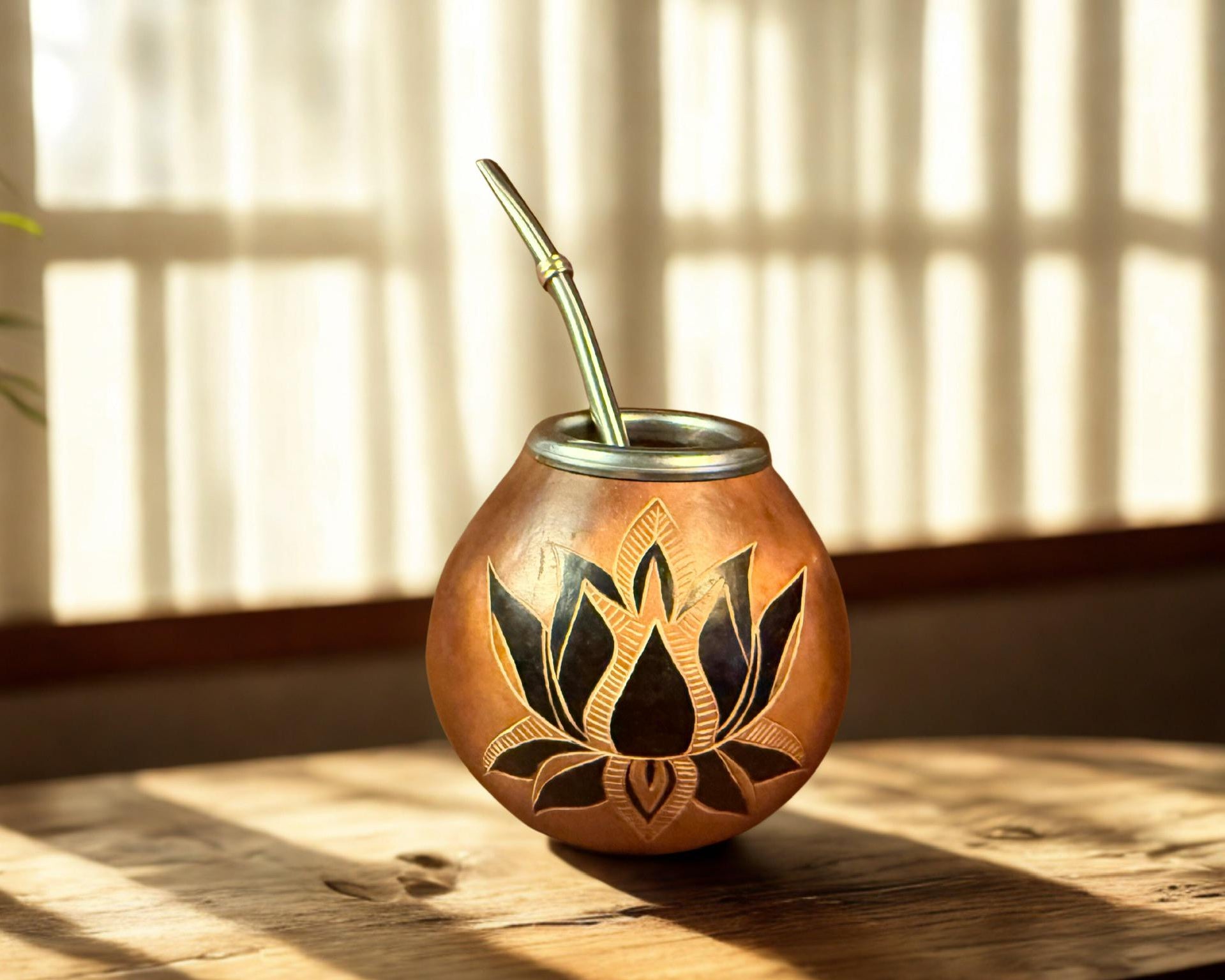 mate cup with a black lotus design and a bamboo straw, set on a wooden table in warm sunlight with window blinds in the background